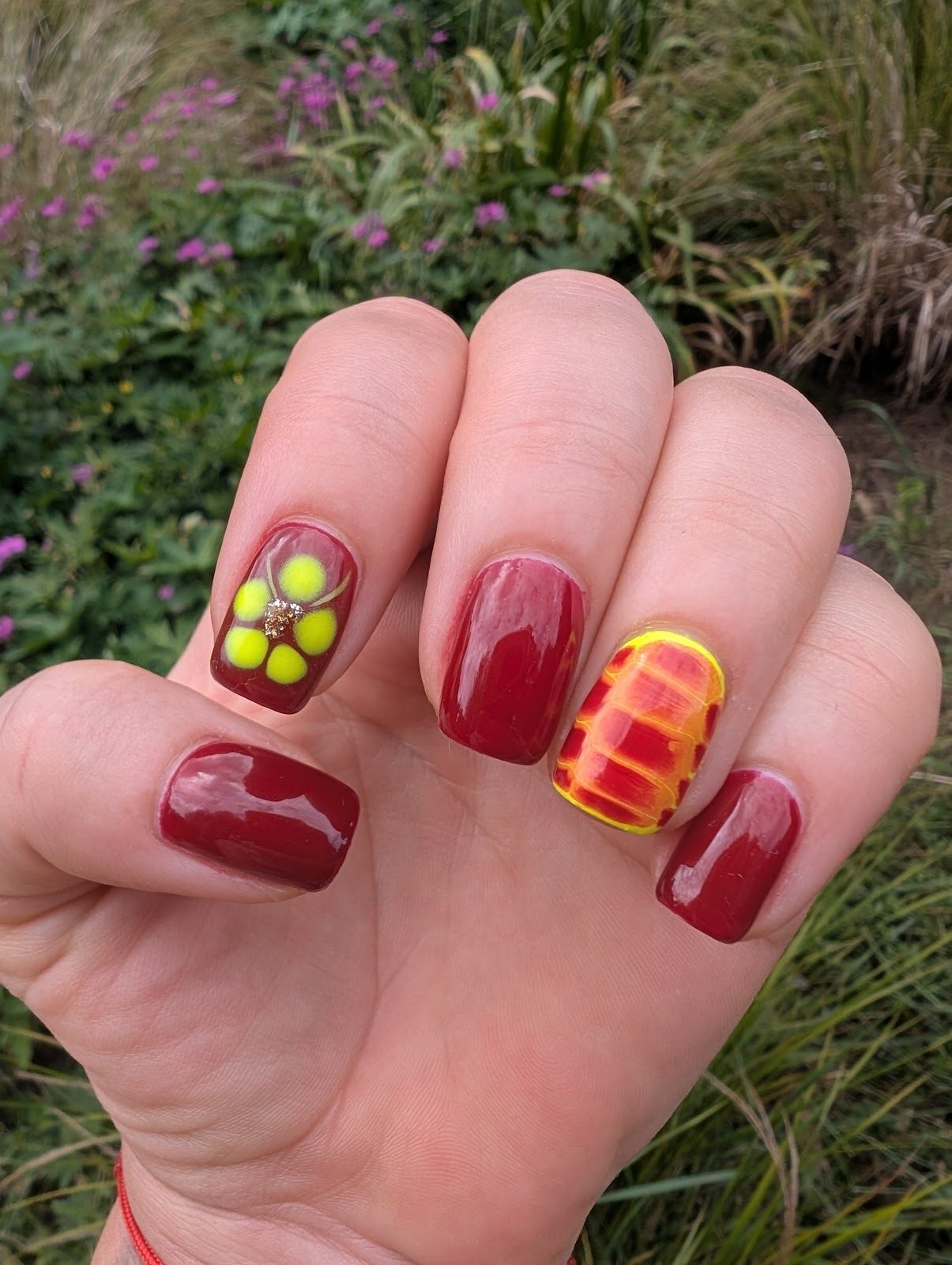 A hand in front of greenery with deep red nails and hand-painted flower and print accent nails