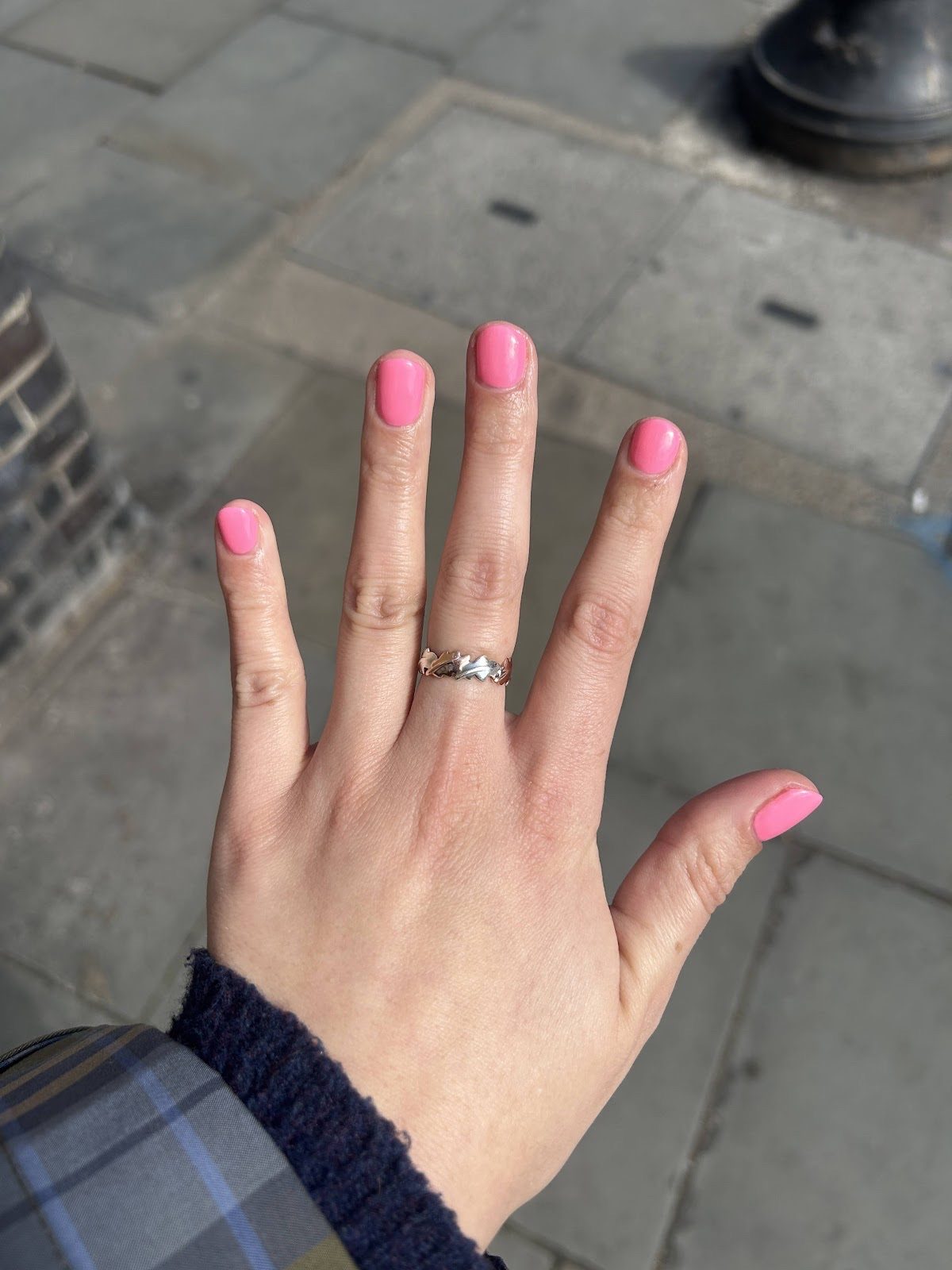 A hand held out over a pavement showing short bright pink nails and a ring